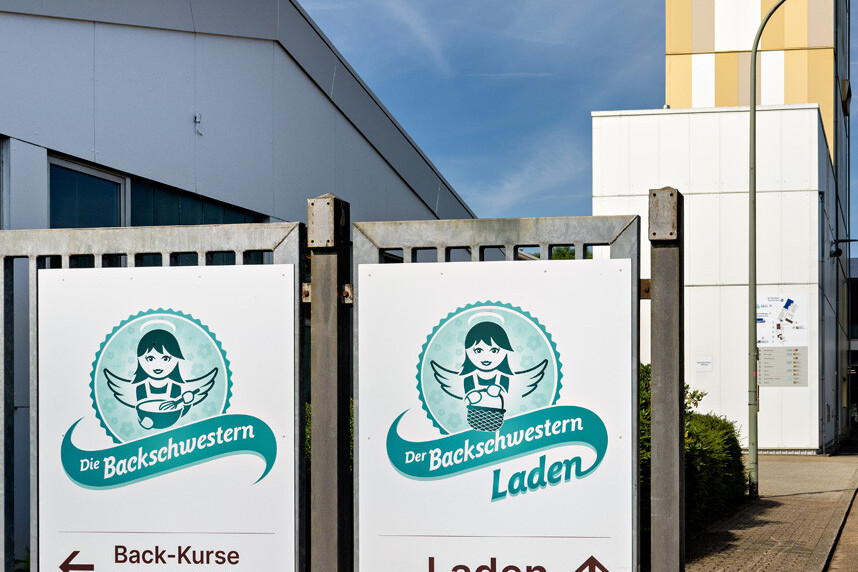 Signposts for “Die Backschwestern” bakery, showing Back-Kurse and Laden directions outside a building.