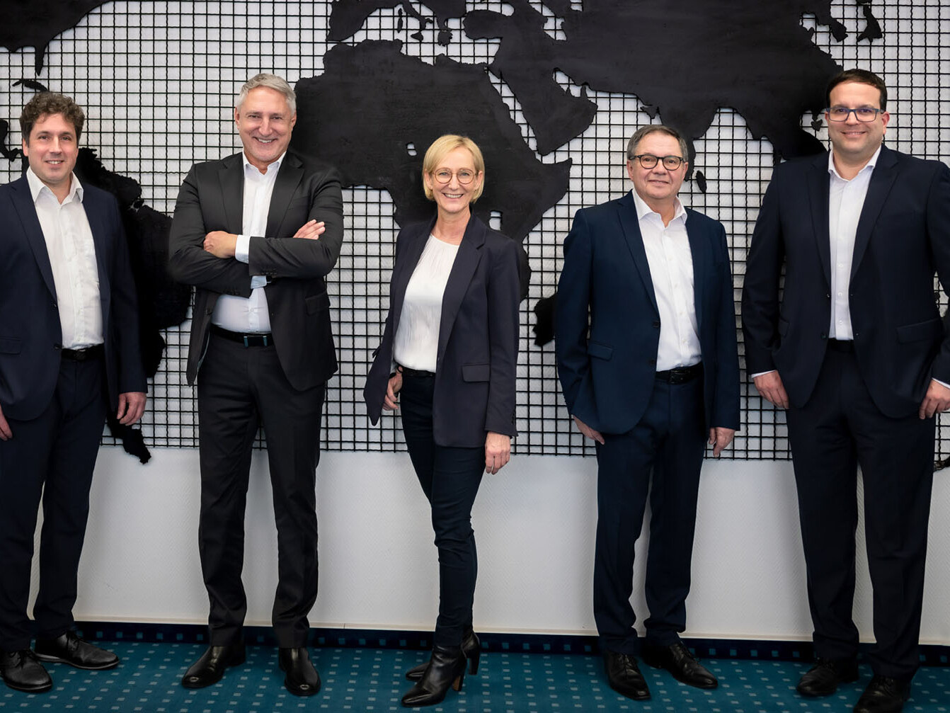 Professional team photo of five executives in suits standing in front of world map backdrop with grid pattern