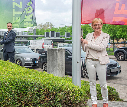 Smiling woman in white blazer posing beside a car at outdoor event with banners and vehicles in background