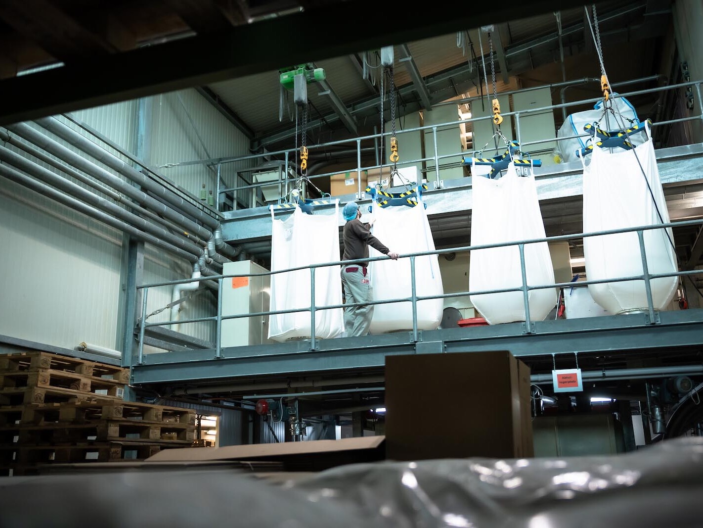 Industrial warehouse worker operating overhead crane lifting large bulk container bags with hooks and slings inside facility