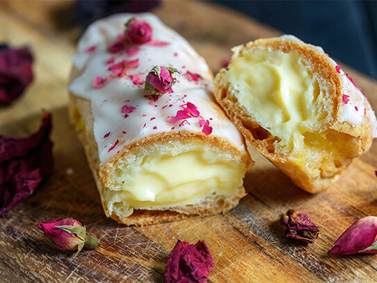 Cream-filled vanilla éclairs with rose petals on rustic wooden board, close-up dessert photo