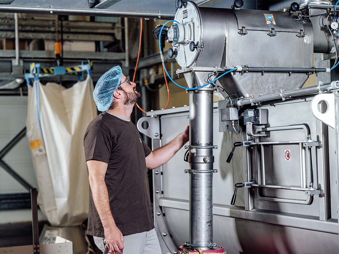 Worker inspecting industrial equipment in a manufacturing facility, wearing safety gear and a blue hairnet.