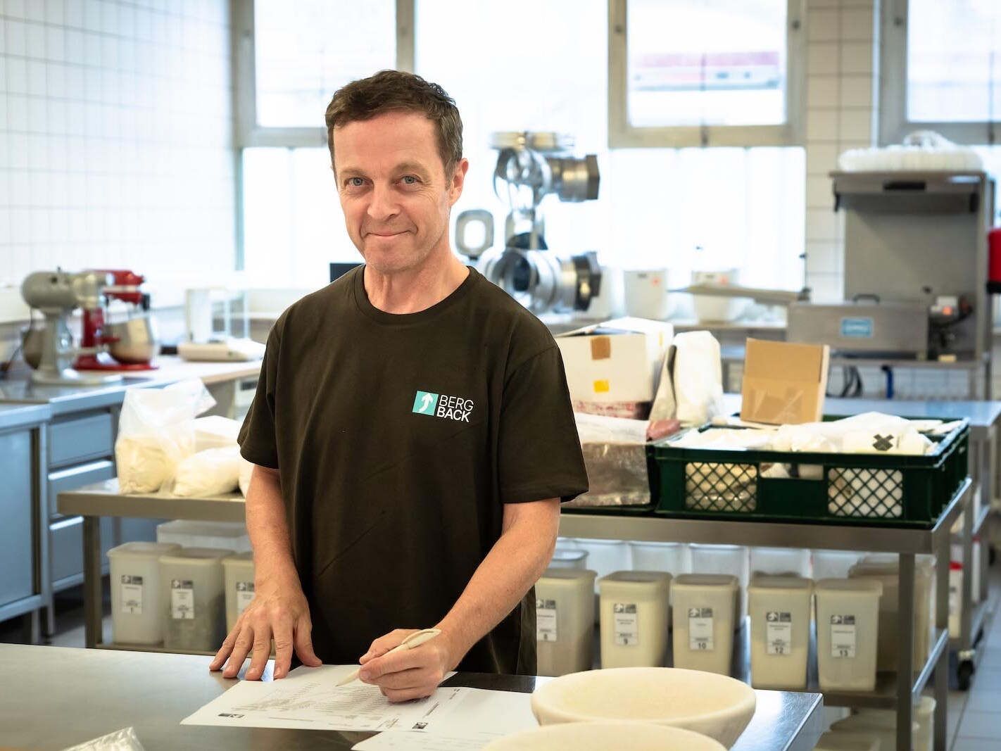 Man in a bakery kitchen wearing a Berg Back shirt, writing on a sheet of paper.