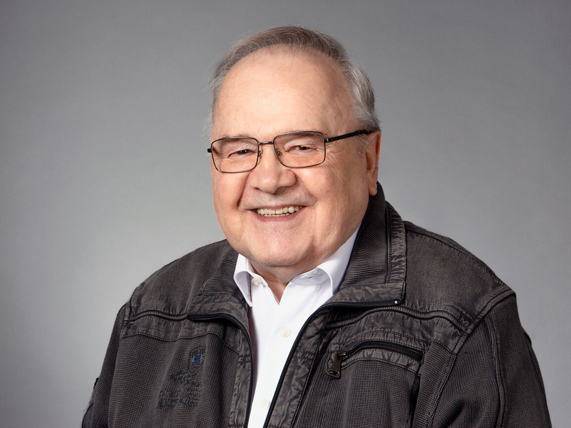 Smiling elderly man in glasses, wearing a gray jacket, against a neutral background.