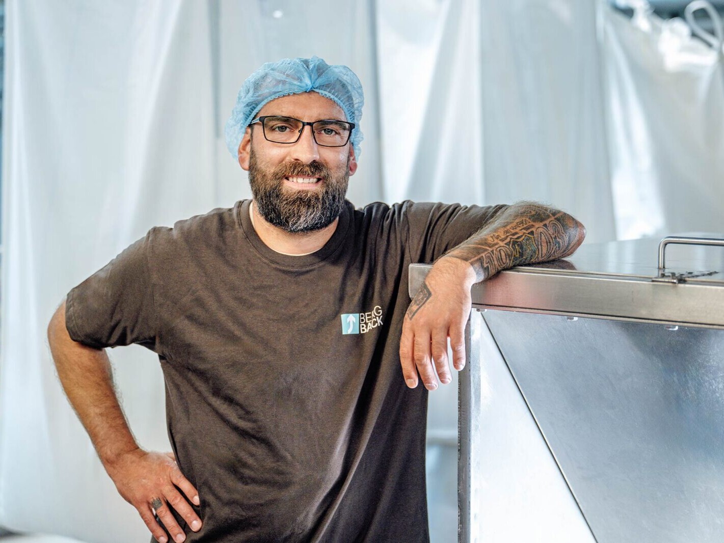 Man with beard and tattoos wearing a hairnet standing in a food production facility, leaning on equipment.