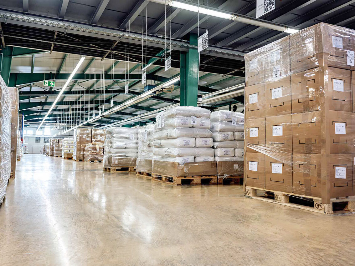 Warehouse interior with stacked pallets of packaged goods and boxes, under high ceilings and fluorescent lights.